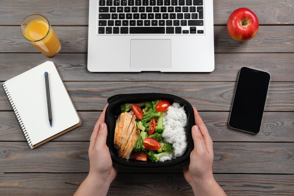 Eine Lunchbox mit Reis, Hähnchen und Gemüse wird vor einem Laptop platziert